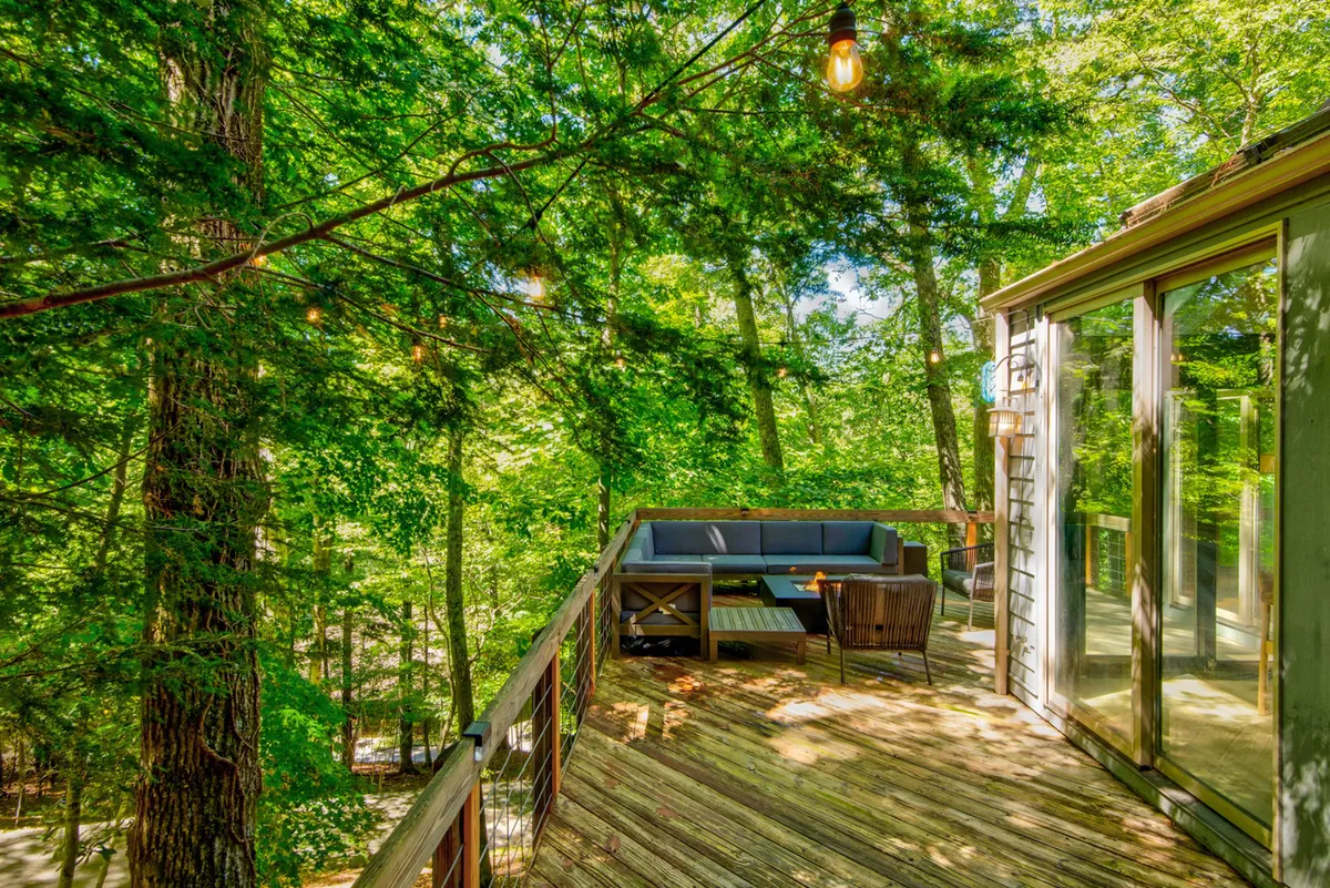 An inviting exterior seating area on a deck, surrounded by tall trees and protected by a railing. The peaceful, sun-dappled setting is perfect for relaxing, reading, or enjoying quality time outdoors, with ambient outdoor lighting for evening enjoyment.
