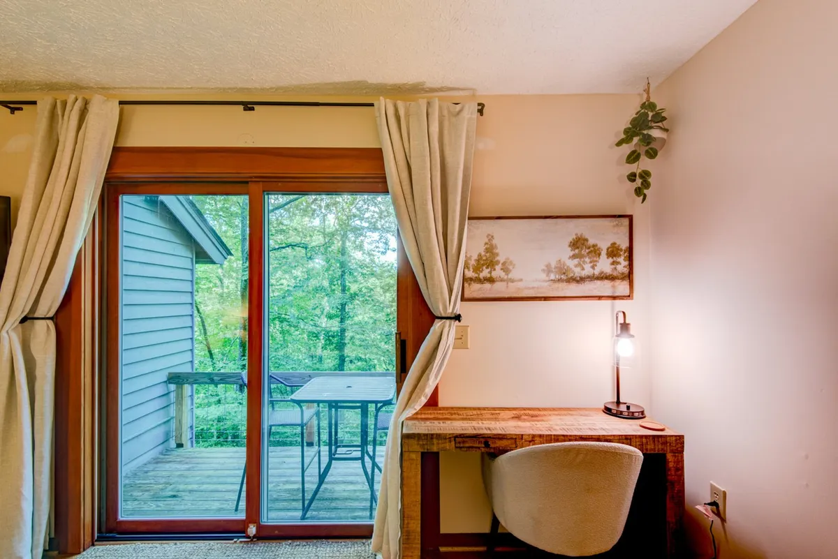 A calm and peaceful space featuring a large glass door leading out to a deck, a wooden desk with a lamp, and decorative wall art. The setting encourages enjoying the view and stepping directly outside to soak in the surroundings.