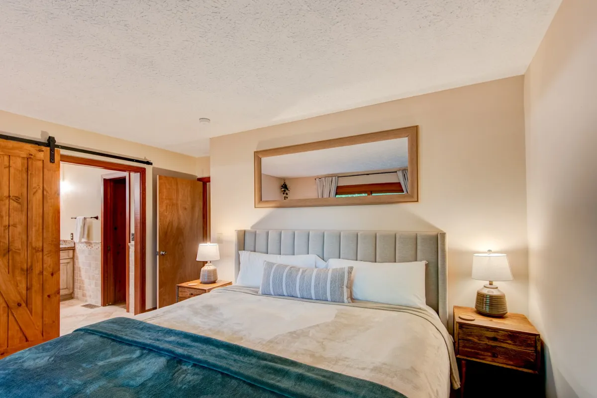 A tranquil bedroom featuring a bed with a gray headboard, decorative pillows, nightstands with lamps, a wall mirror, and a wooden sliding door. The space exudes comfort, offering a peaceful retreat for rest or quiet moments.