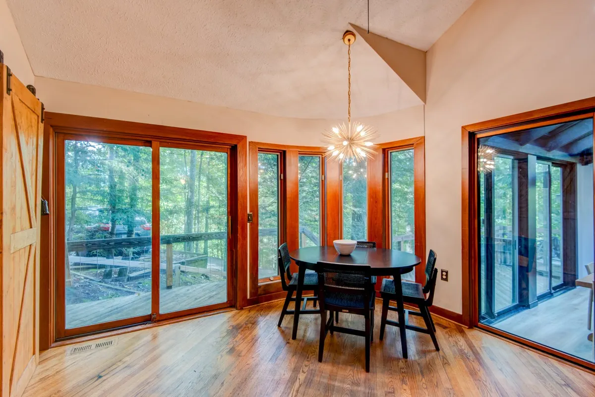A dining table is set near floor-to-ceiling windows, flooding the space with natural light and offering views of the surrounding landscape. The unique light fixture and the visual connection to the outdoors make this space ideal for enjoying meals while immersed in nature.