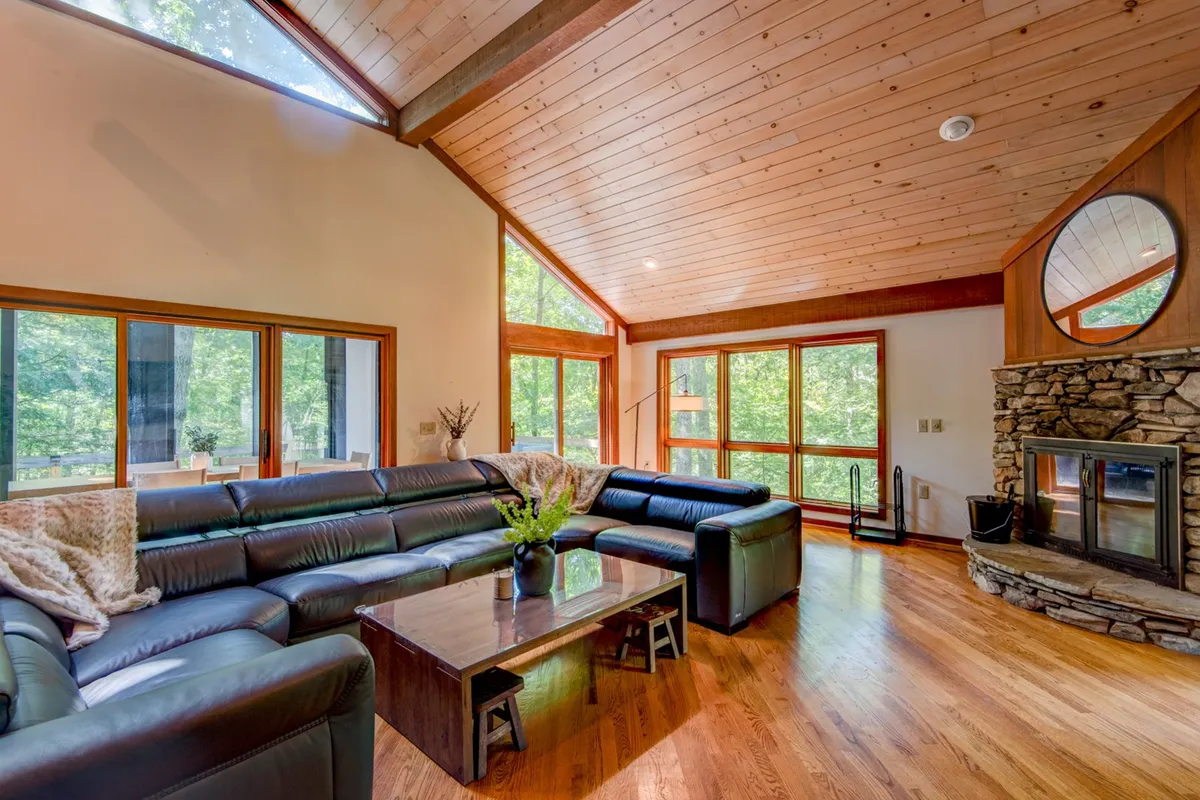 This image features a comforting living space with a leather sectional sofa, a striking stone fireplace, and expansive large windows that frame the natural landscape outside. The wooden ceiling adds to the rustic, mountain-lodge ambiance, and the windows bring the outdoors into the room, making nature an integral part of the experience.