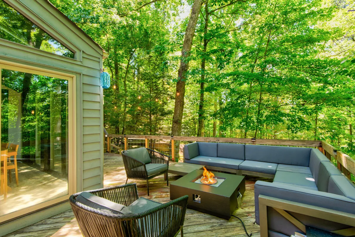 This image showcases an inviting outdoor seating area featuring a cushioned sectional and a gas fire-table, surrounded by mature trees. The setting is perfect for social gatherings, relaxing evenings, or outdoor dining, offering a cozy and communal ambiance that matches the section's narrative of relaxation and togetherness.