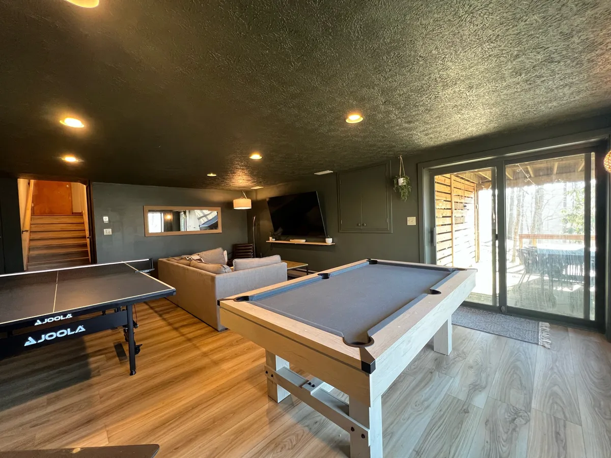 This lively interior scene showcases a game room equipped with both a ping-pong table and pool table, with a comfortable sectional sofa seating area and a sliding glass door that leads outside. The arrangement encourages friendly competition and socialization, while the proximity to the outdoors hints at seamless transitions between indoor and al fresco fun.
