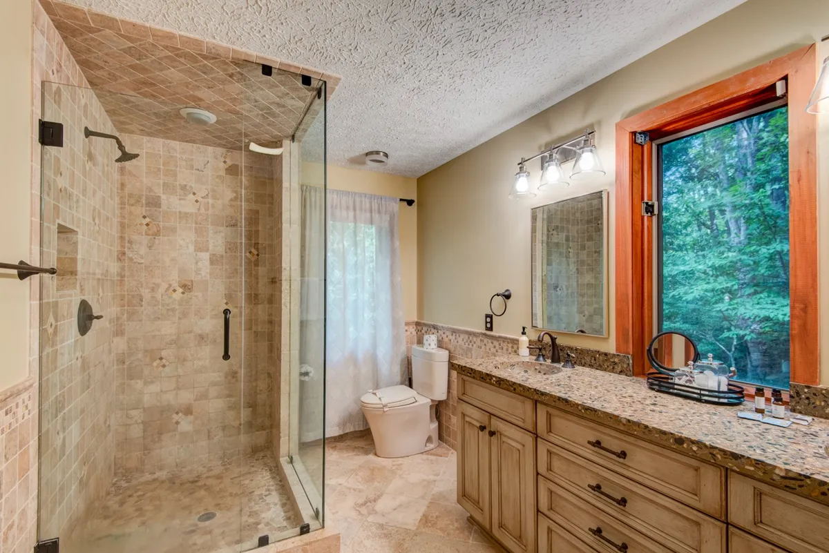 A spacious bathroom with a glass shower enclosure, granite countertop, large window offering a forest view, and a double vanity. The area is bathed in natural light, creating a spa-like, rejuvenating environment.