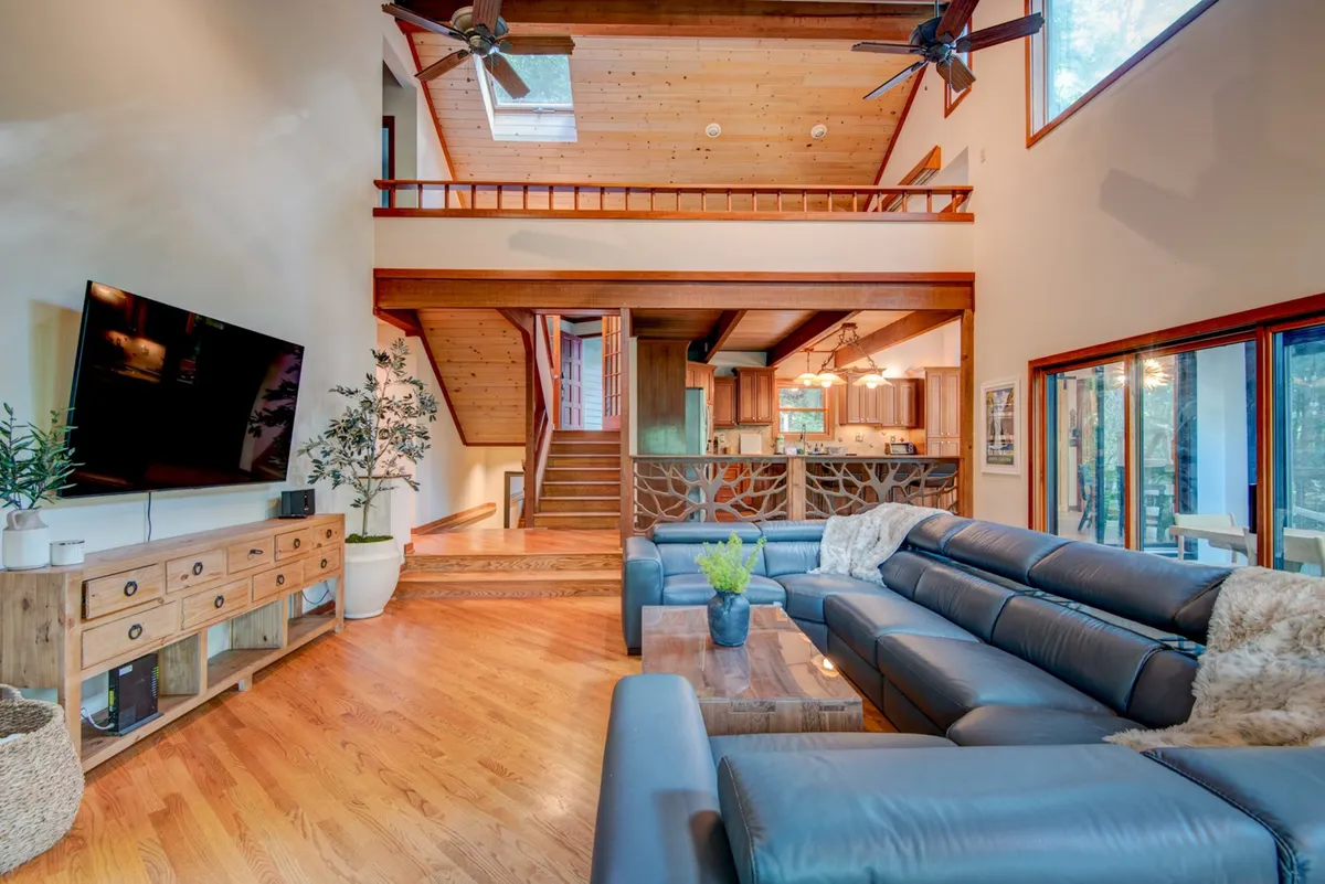 This image features a spacious living area with a large sectional sofa, wooden coffee table, TV mounted on the wall, and a staircase leading to the upper level. The area is flooded with natural light from large windows, creating a warm and inviting atmosphere ideal for gatherings and relaxation after outdoor adventures.