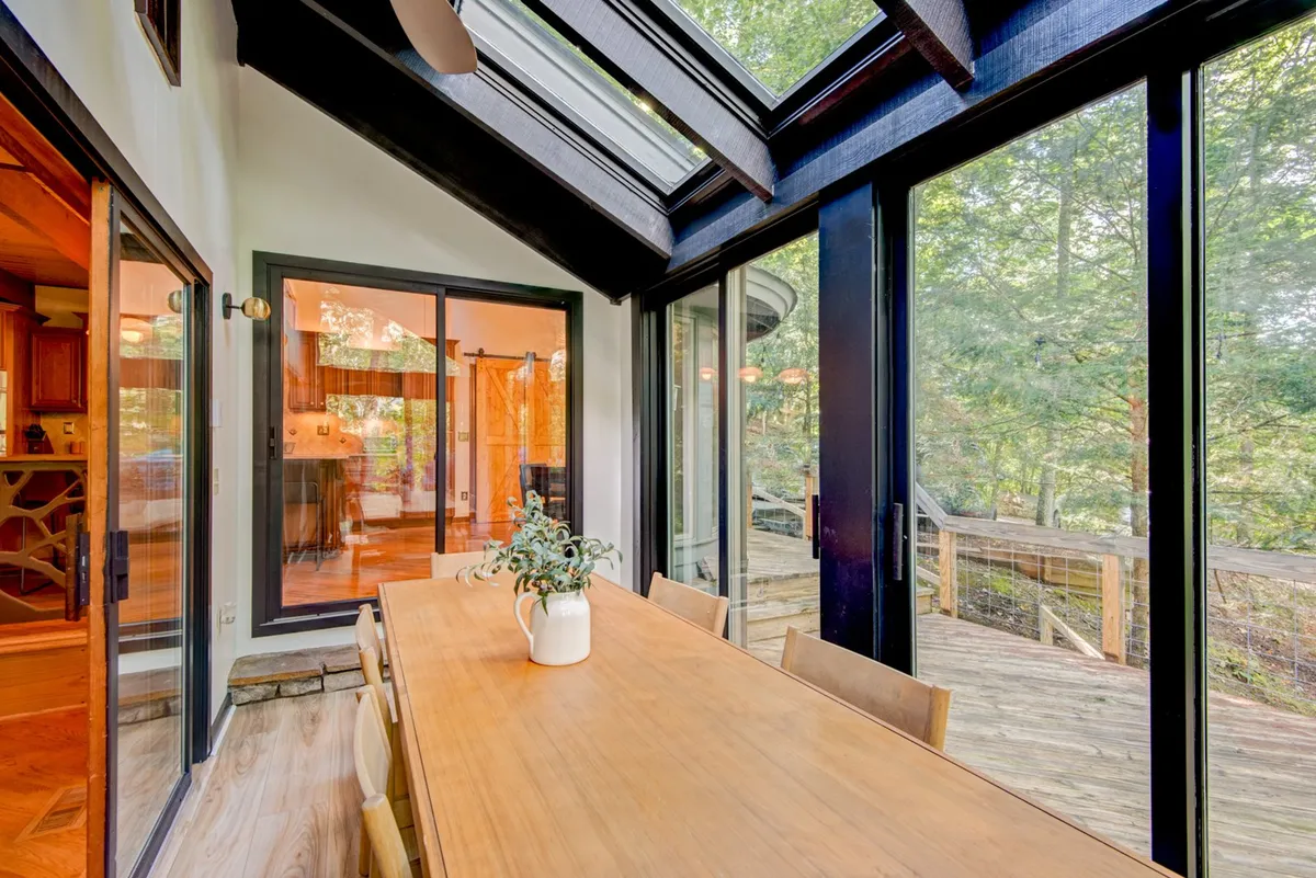 A sunlit dining room with a large table set beneath skylights. Surrounded by glass walls, the space provides sweeping views of the greenery outside, making every meal feel connected to nature and full of light.