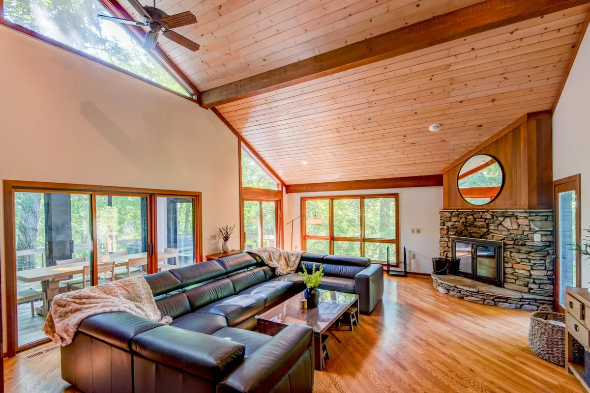 A cozy living room scene featuring a plush leather sectional sofa centered around a striking stone fireplace. Large windows flood the space with natural light and offer views of the surrounding greenery, while the inviting layout encourages gathering and relaxation.
