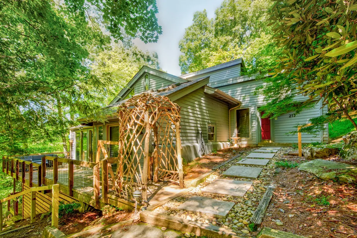 The image showcases the inviting front entrance of Deerwood Manor, featuring a charming decorative trellis, a winding stone pathway, and lush greenery that frames the home. The peaceful mountain setting is evident, with mature trees and tranquil landscaping hinting at the serene retreat that awaits guests.