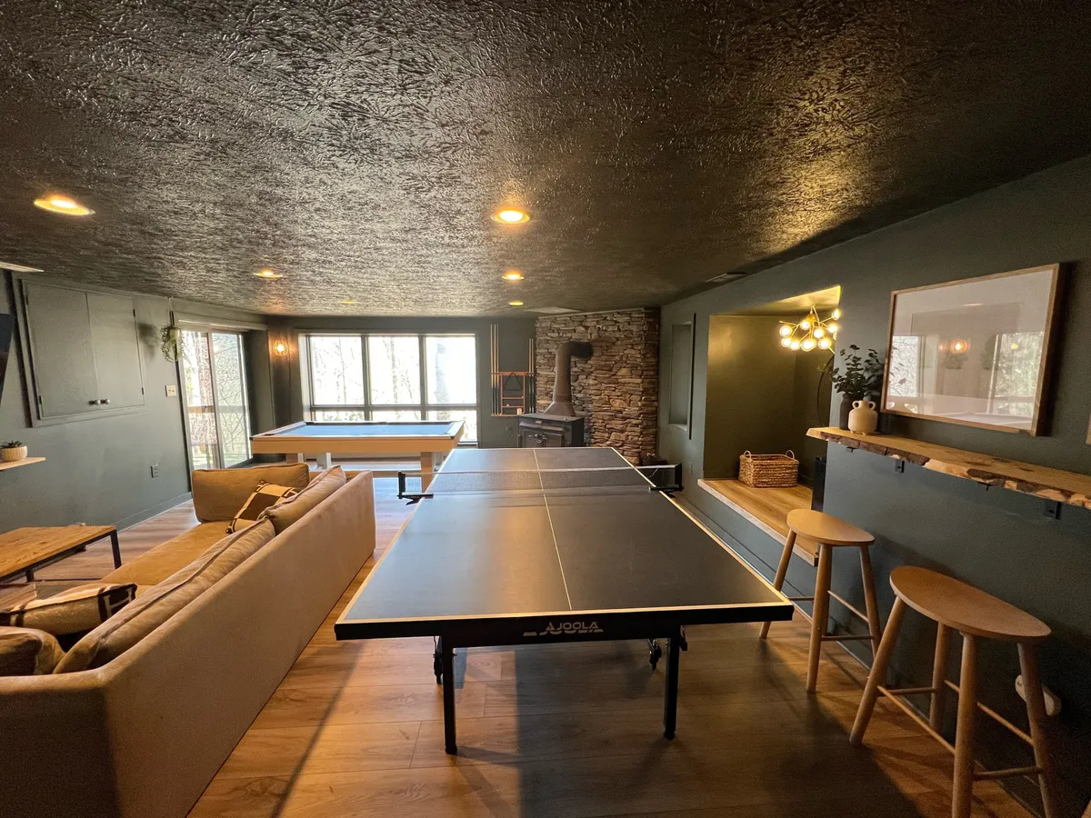 A vibrant lower-level entertainment space featuring a ping-pong table, plush sofa, expansive windows letting in natural light, and a cozy stone fireplace. The scene exudes a welcoming, energetic vibe, ideal for both spirited game nights and relaxed gatherings by the fire.