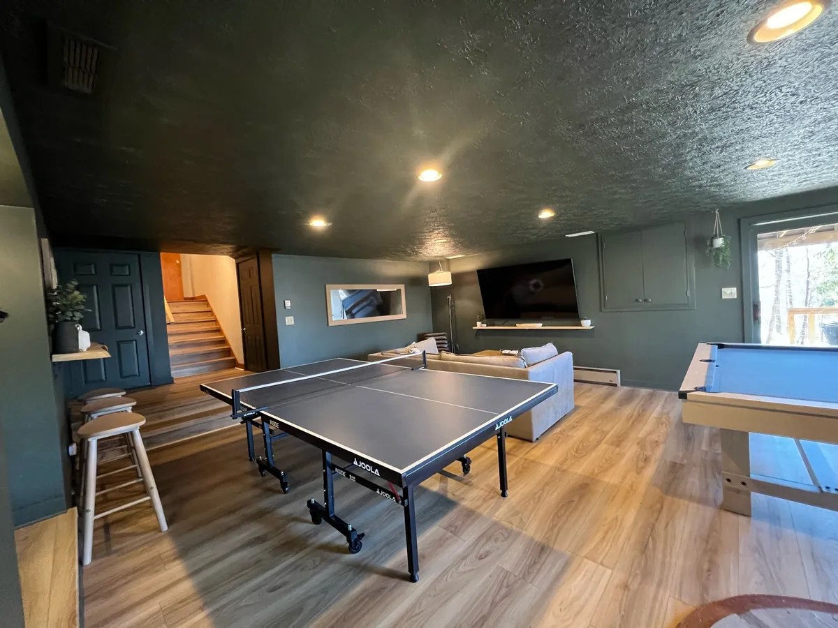 This image showcases a ping pong table as the focal point, with a sofa, television, and staircase in the background. The space is clearly designed for social interaction, friendly competition, and energetic gatherings — all in a casual, inviting environment.