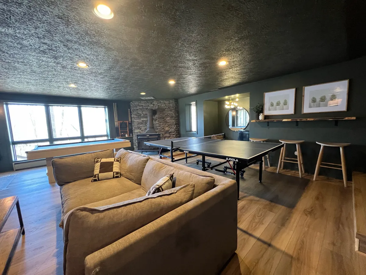 A lively interior space featuring a comfortable sofa, large windows with ample natural light, a stone fireplace, and a ping pong table. The room is designed for both relaxation and play, suitable for guests of all ages.