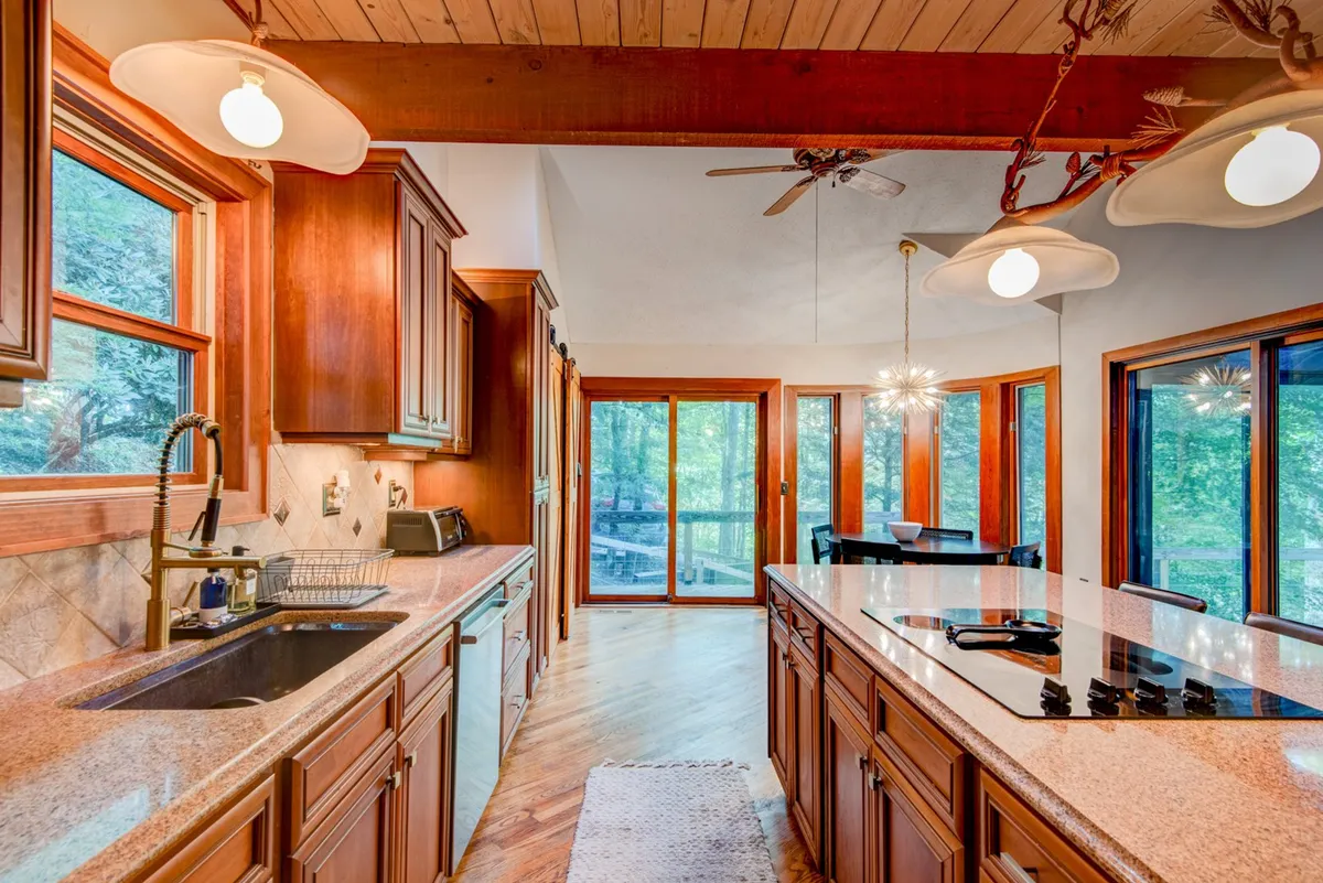 A warm, inviting kitchen scene showcasing granite countertops, wooden cabinetry, a pantry door, and large windows that let in ample sunlight. The open layout includes direct views to an adjacent dining area, encouraging social interaction and shared culinary experiences.